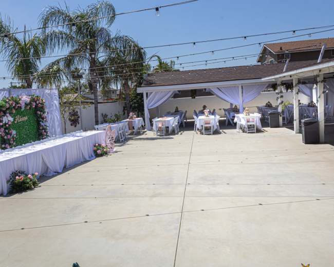 The image shows a spacious outdoor venue set up for an event, featuring decorated tables and a floral backdrop, with palm trees and string lights overhead.