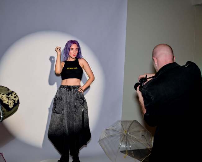 A model with purple hair poses in a black crop top and cargo pants while a photographer captures the scene.