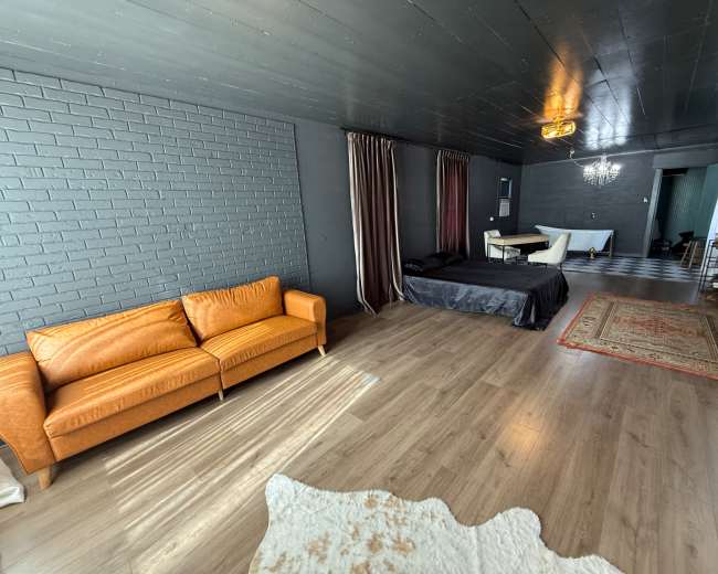 The room features dark walls, a wooden floor, and a mix of furniture including an orange sofa, a black bedspread, and a patterned rug.
