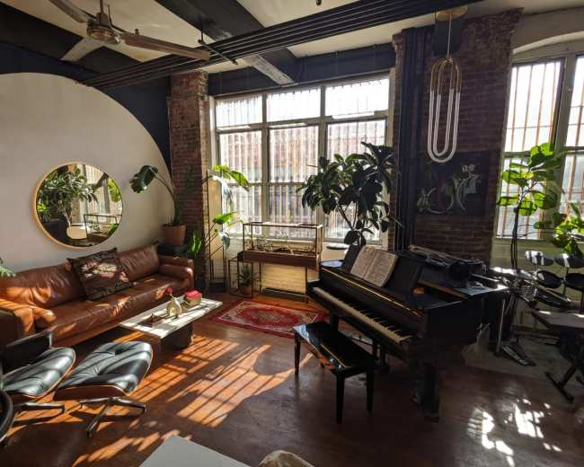 The image shows a cozy living room featuring a grand piano, a brown leather sofa, a round mirror, several indoor plants, and large windows allowing light to illuminate the space.