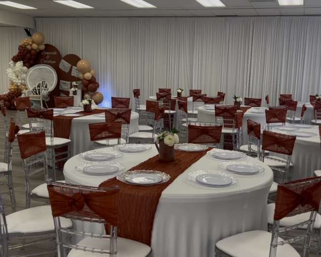 The image shows a banquet hall set up for an event, with round tables covered in white tablecloths and adorned with orange ribbons, alongside decorated walls and a backdrop featuring balloons.