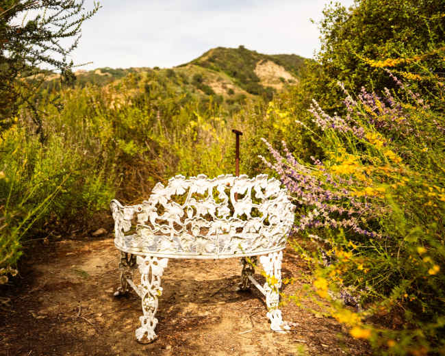 A decorative white bench sits amidst lush greenery and wildflowers, with rolling hills in the background.