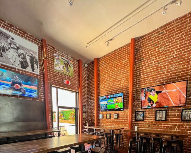 The interior of a sports bar features exposed brick walls, wooden tables, and two televisions displaying sporting events.