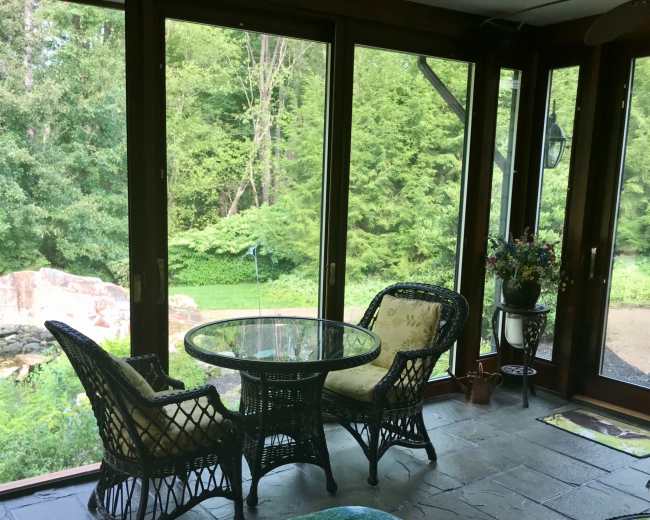 A sunroom features two wicker chairs and a glass table, overlooking a lush garden through large windows.