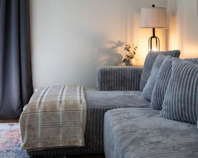 The image shows a gray, corded sectional sofa with decorative cushions and a throw blanket, paired with a floor lamp and a plant on a side table.