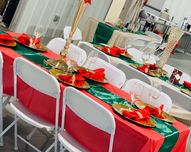 A decorated dining room with tables covered in red and green cloth, featuring gold candlesticks, plates, and napkins arranged for a festive event.