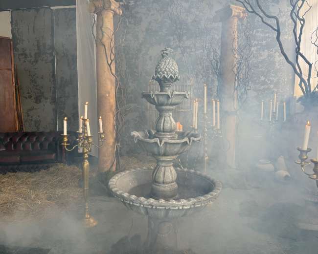 A decorative stone fountain with a pineapple sculpture sits in a foggy room surrounded by candelabras and overgrown greenery.