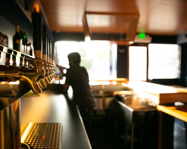 A silhouette of a person stands near a bar with several beer taps in a dimly lit space.
