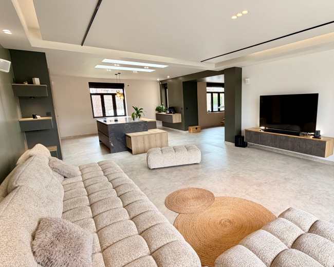 The image depicts a spacious living room featuring a large, light-colored sectional sofa, a circular area rug, and a mounted television with a wooden media console against a neutral wall.
