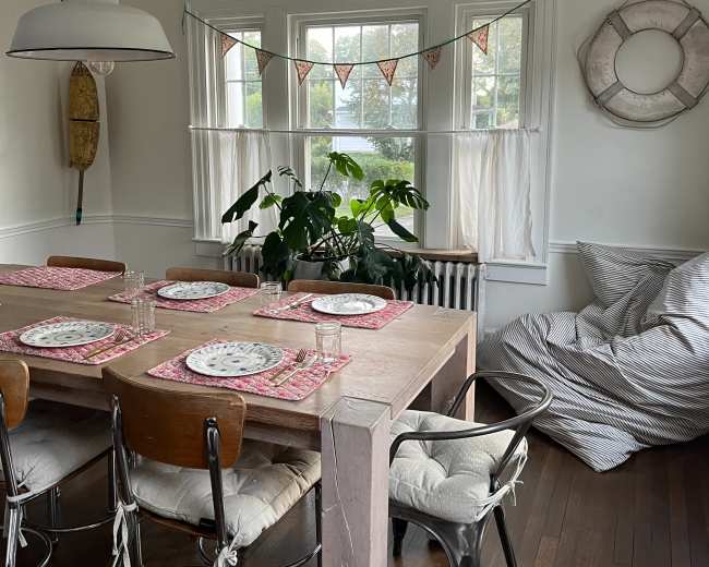A dining room features a wooden table set with plates and glasses, surrounded by chairs, with a large plant by the window and a cozy seating area in the corner.