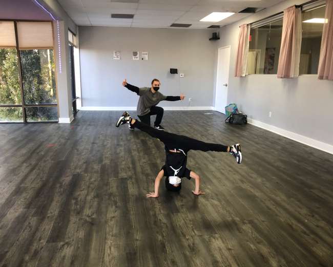 Two individuals are performing dance poses in a spacious, brightly lit studio with wooden flooring.