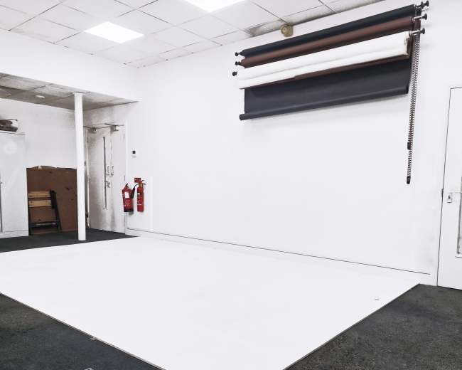 The image shows an empty studio space with a white wall and a roll of backdrop paper mounted on the wall, alongside fire extinguishers in the corner.
