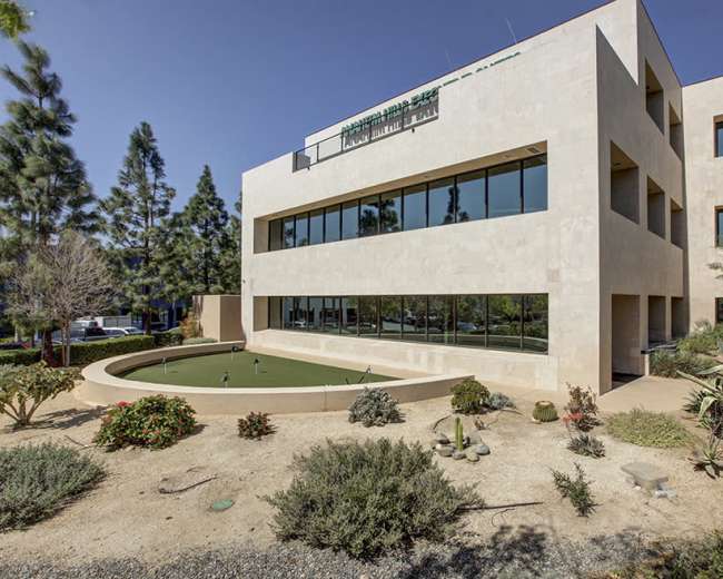 The image shows a modern, multi-story commercial building with a fountain and landscaped gardens in front.