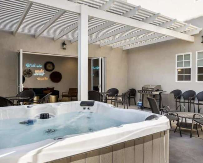 A hot tub is situated under a pergola in an outdoor seating area with chairs and a grill nearby.