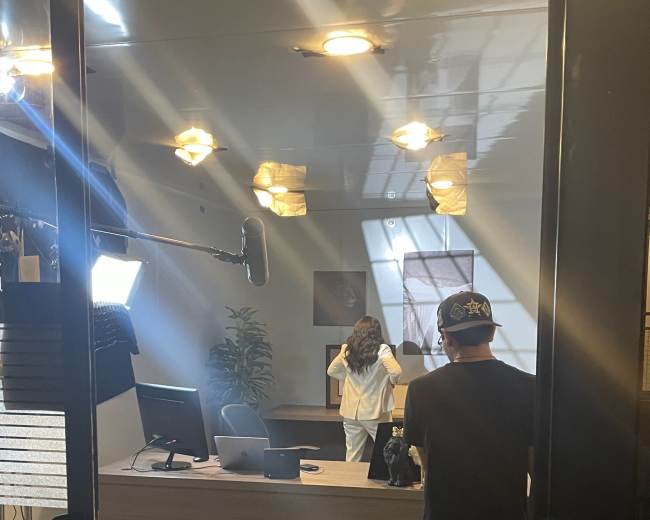 A person stands in an office setting while a camera operator captures the scene.