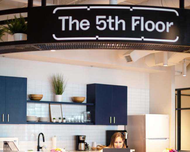 A woman works on a laptop at a modern reception desk in an office space labeled "The 5th Floor."