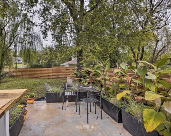 A backyard terrace features a dining set surrounded by planters and a hammock under the trees.