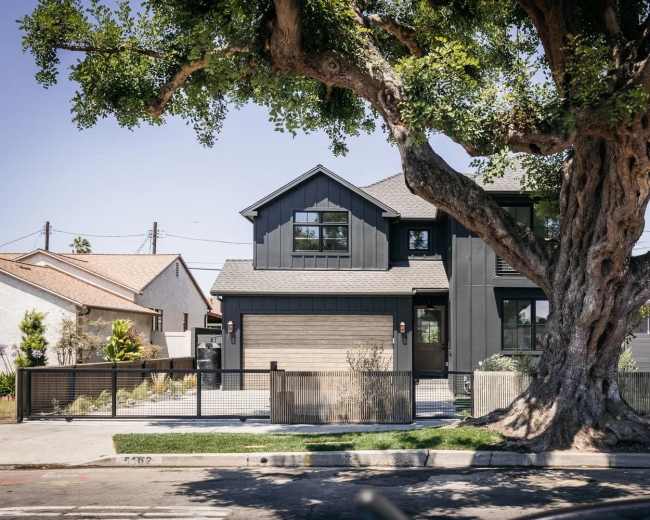 A modern two-story house with a dark exterior and large windows stands next to a massive tree, surrounded by a fence and well-kept landscaping.