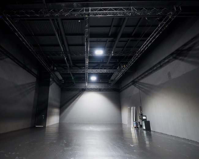 The image shows a large, empty studio space with dark walls, a polished floor, and industrial lighting overhead.