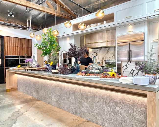 A modern kitchen displays a large, textured countertop adorned with an array of colorful food items and elegant serving dishes, while a person prepares in the background.