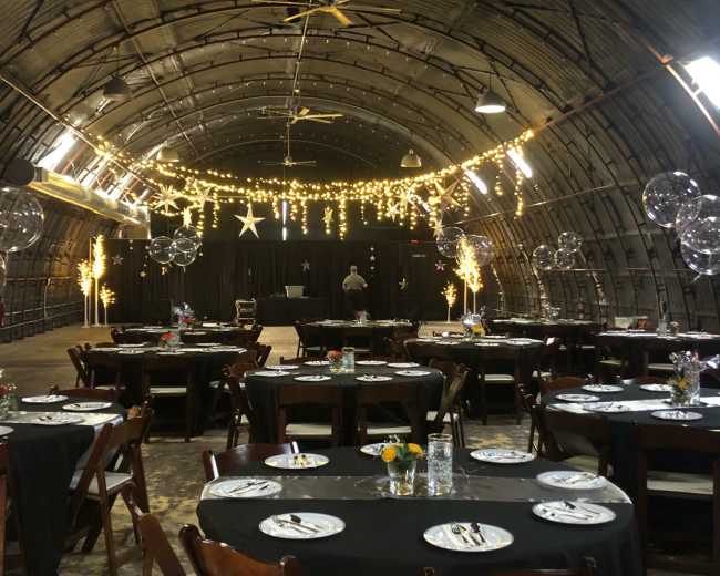 The image shows an elegantly arranged dining area inside a large, curved space, with tables set up for an event and decorative lights hanging from the ceiling.