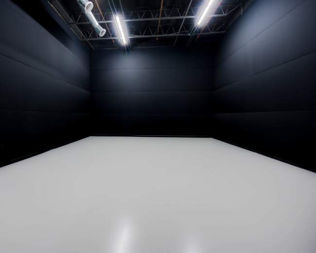 A large, empty room with black walls and a white floor, illuminated by overhead lights.