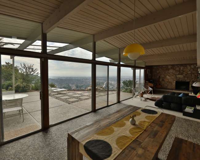 The interior of a modern house features large glass doors opening to a patio with a view overlooking a cityscape, and a dining area with a wooden table and distinct decor.