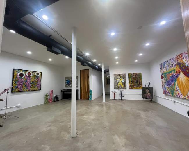 The image shows a spacious art studio or gallery with several colorful paintings on the walls, a keyboard, and various art supplies scattered around the room.