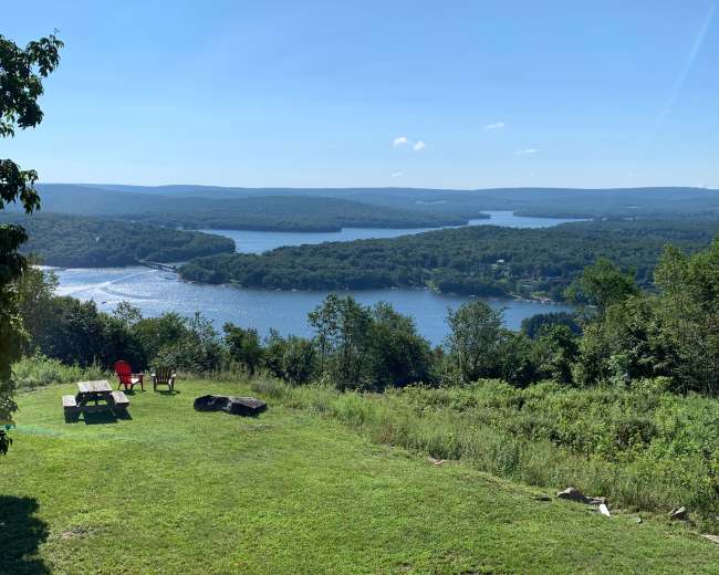 The image shows a grassy hillside overlooking a river surrounded by green hills and trees under a clear blue sky.