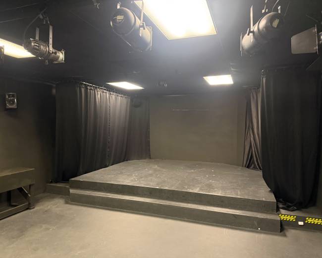The image shows a small black box theater with a raised stage, black curtains, and overhead lighting fixtures.