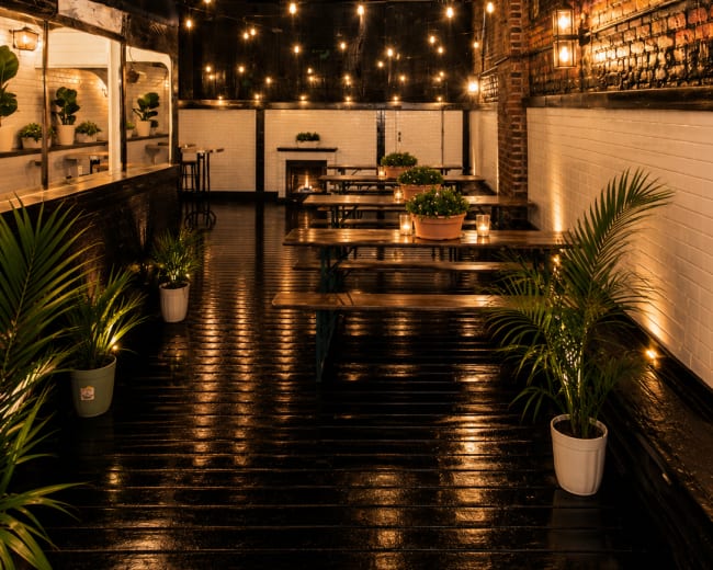 The image shows a rooftop outdoor dining area with wooden tables, potted plants, and string lights, surrounded by brick walls and a mural of a city skyline.