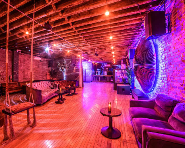 The image shows an interior of a rustic lounge with exposed wooden beams, plush seating, and colorful lighting effects.