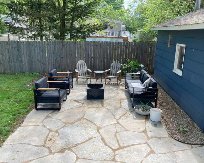 A patio area with a circular seating arrangement around a fire pit, surrounded by a wooden fence and greenery.