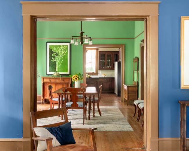 The image shows a brightly colored room with blue walls and a green room visible through an open doorway, featuring wooden furniture and a dining area.