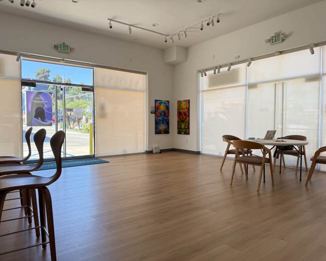 The image shows a bright, minimalist interior of a room with wooden flooring, a table and chairs set up, and large windows letting in natural light.