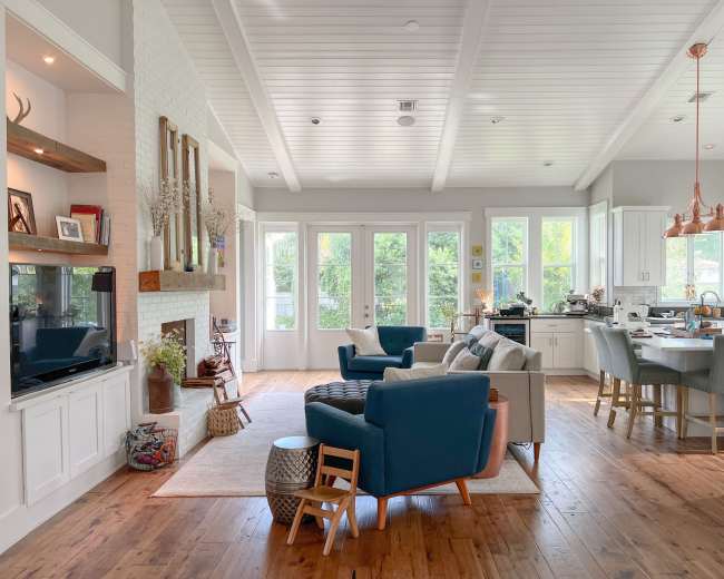 The image shows a bright, open living area with a blue and beige seating arrangement, a dining space with wooden chairs, and large windows that provide natural light.