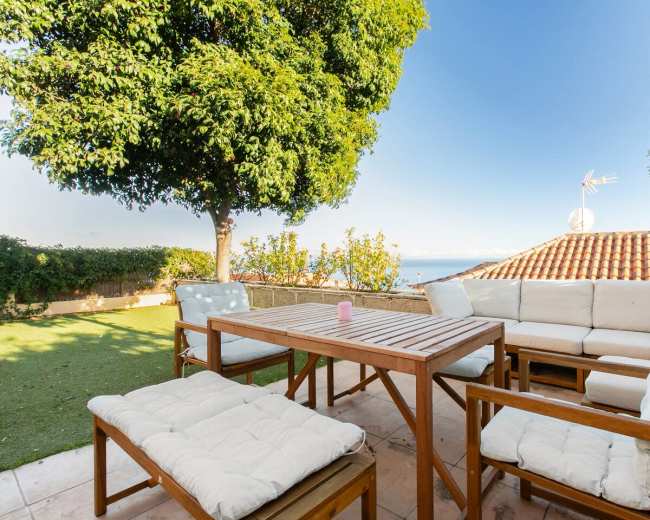 A patio with wooden furniture and cushions overlooks a green lawn and a distant view of the ocean under a clear sky.