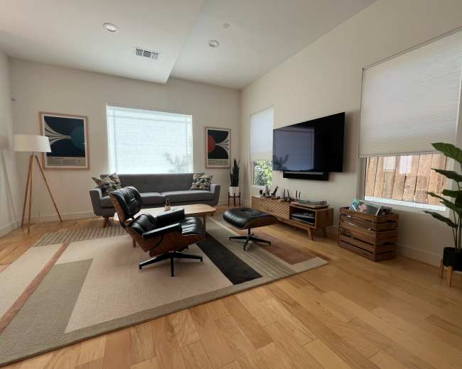 The image shows a modern living room featuring a gray sofa, a black lounge chair with an ottoman, a flat-screen television mounted on the wall, and wooden flooring.