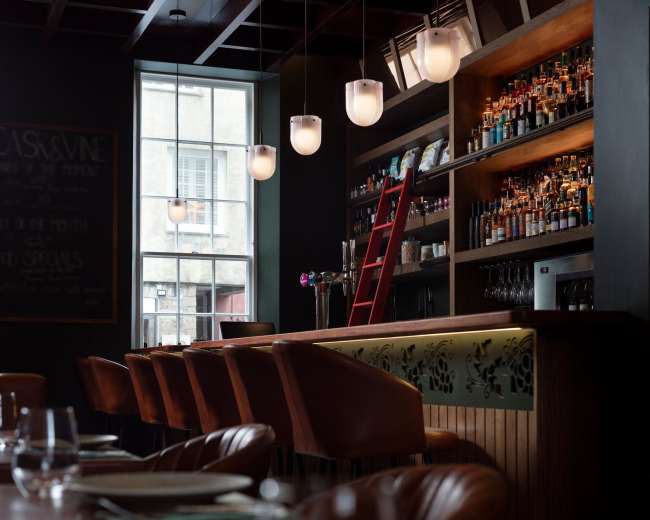 The image shows a dimly lit bar area with wooden shelves filled with various bottles, a red ladder leaning against the shelf, and a large window letting in natural light.
