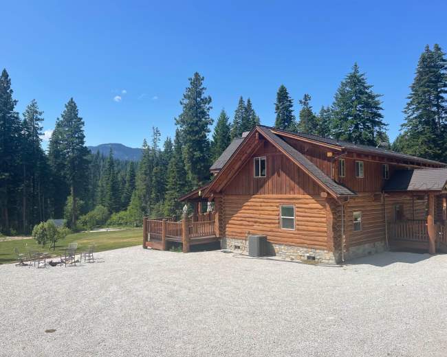 A large log cabin is surrounded by dense trees and a gravel area, with mountains visible in the background.