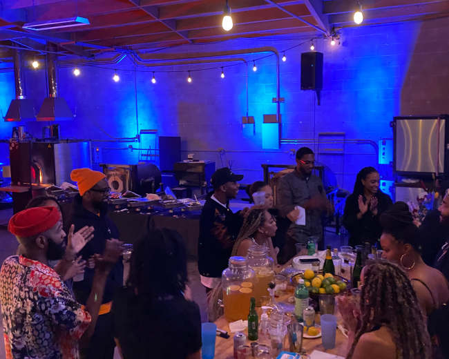 A group of people gathers around a large table filled with drinks and food in a stylish, well-lit space with industrial decor.