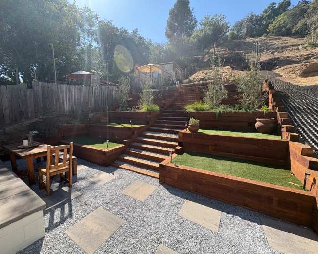 The image shows a landscaped backyard with terraced wooden planters, stone steps leading up, and a table and chairs in a gravel area.