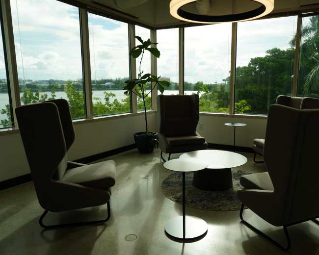 The image shows a modern seating area with four high-backed chairs and a low coffee table, surrounded by large windows that provide a view of greenery and water outside.