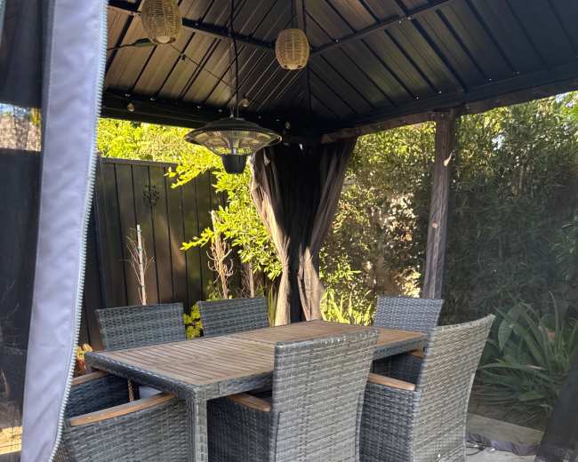 A set of six wicker dining chairs surrounds a rectangular table under a gazebo with greenery in the background.
