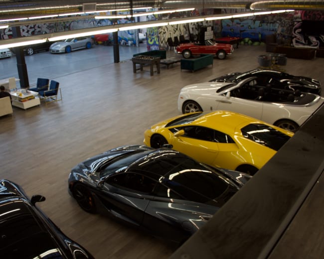 The image shows a spacious garage featuring several luxury cars, including a yellow sports car and a white coupe, with two people seated at a table in the background.