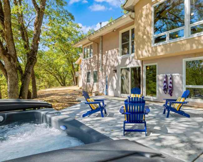 A patio with four blue chairs surrounds a hot tub near a house with large windows and trees in the background.