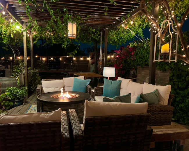 A cozy outdoor seating area features a circular fire pit surrounded by wicker chairs with light blue cushions, illuminated by string lights and lanterns.