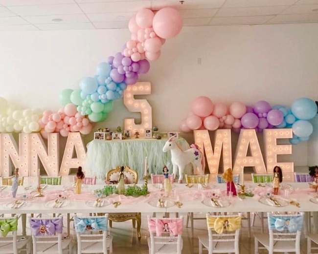 A children's birthday party setup features a decorated table with pastel balloons, a large "JENNA" and "5" sign, and various themed decorations.