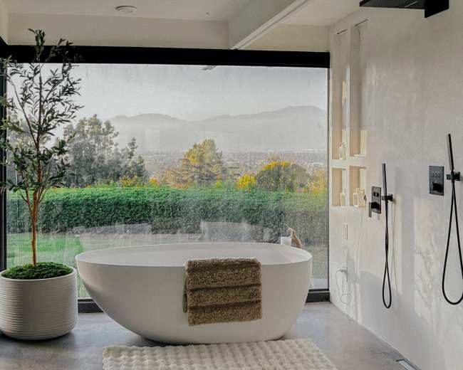 The image shows a modern bathroom with a freestanding bathtub, rainfall shower fixtures, and a large window overlooking a scenic view of mountains and greenery.