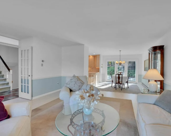 The image shows a bright living room with two sofas, a glass coffee table, and a dining area visible through a set of glass doors.